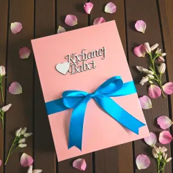 Zamknięte różowe pudełko przewiązane niebieską wstążką z drewnianym napisem „Kochanej Babci”